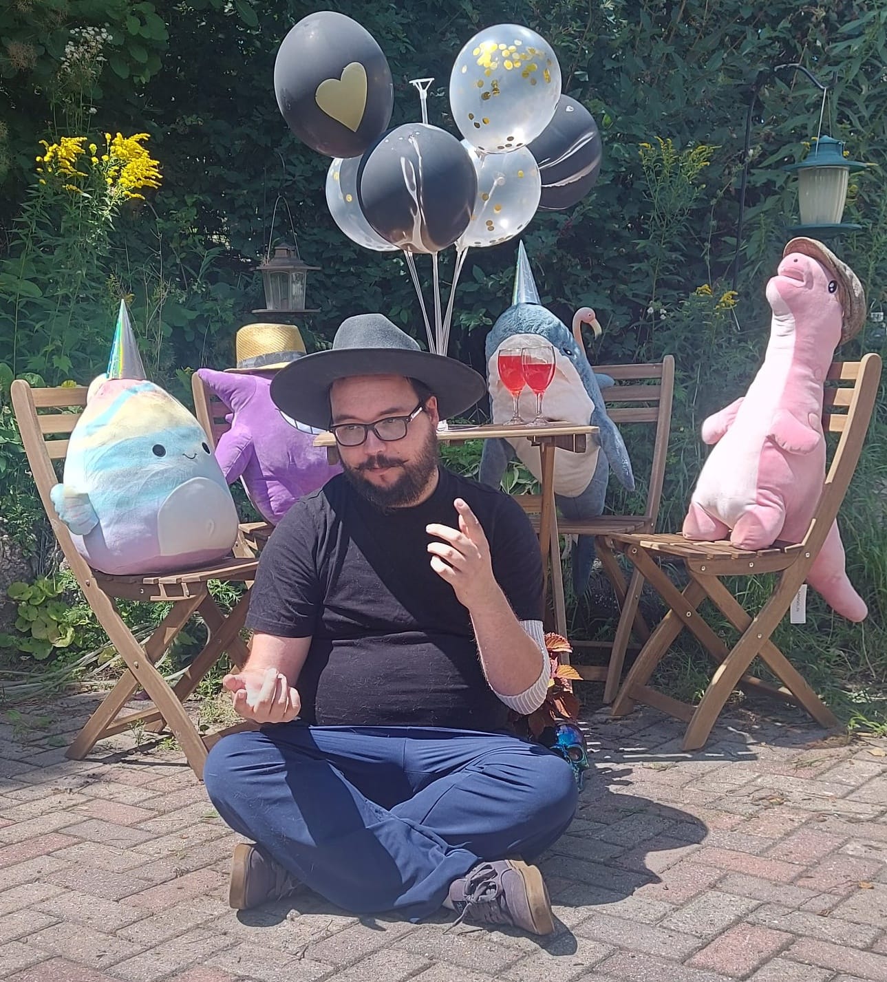 A man with facial hair and a felt sun hat sits on the ground in front of a patio set hosting a stuffed animal patio party, Photo 3